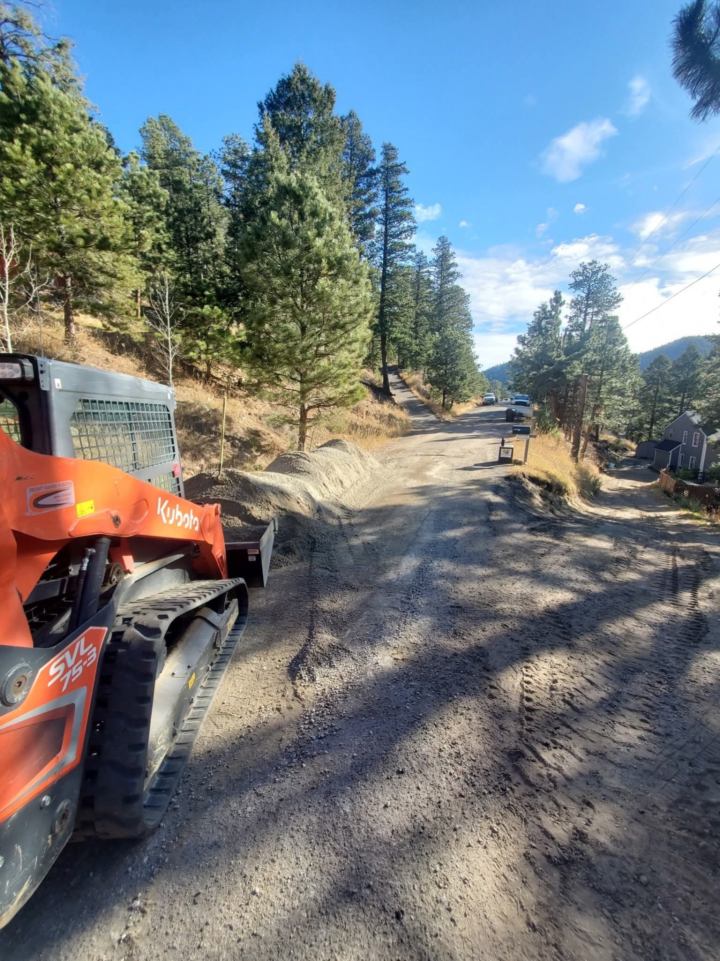 Orange Kubota SVL75-3 skid steer parked on a mountain road next to a gravel pile, with pine trees and a clear blue sky.