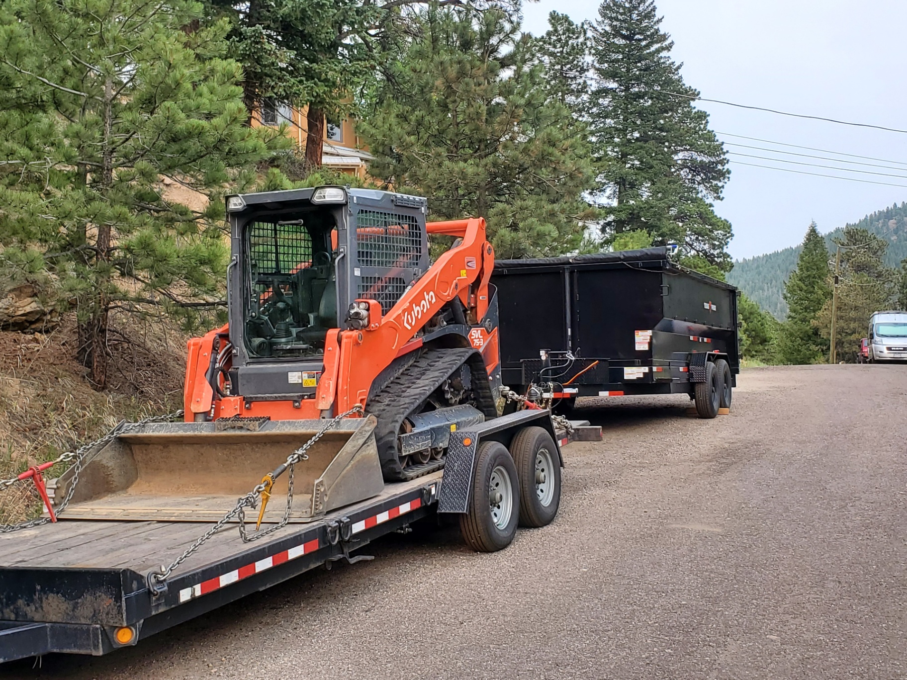 Move Earth rig on an Evergreen mountain road: white Ford truck towing an orange Kubota SVL75-3 skid steer on a flatbed with a black dump trailer behind