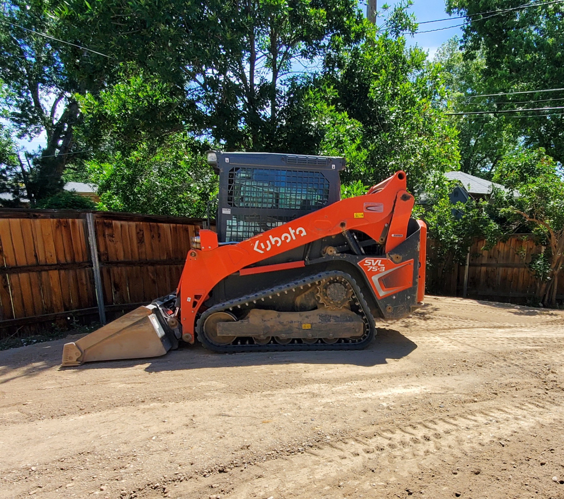 Orange Kubota SVL 75-3 compact track loader parked on a dirt lot with bucket lowered, wooden fence and trees behind it