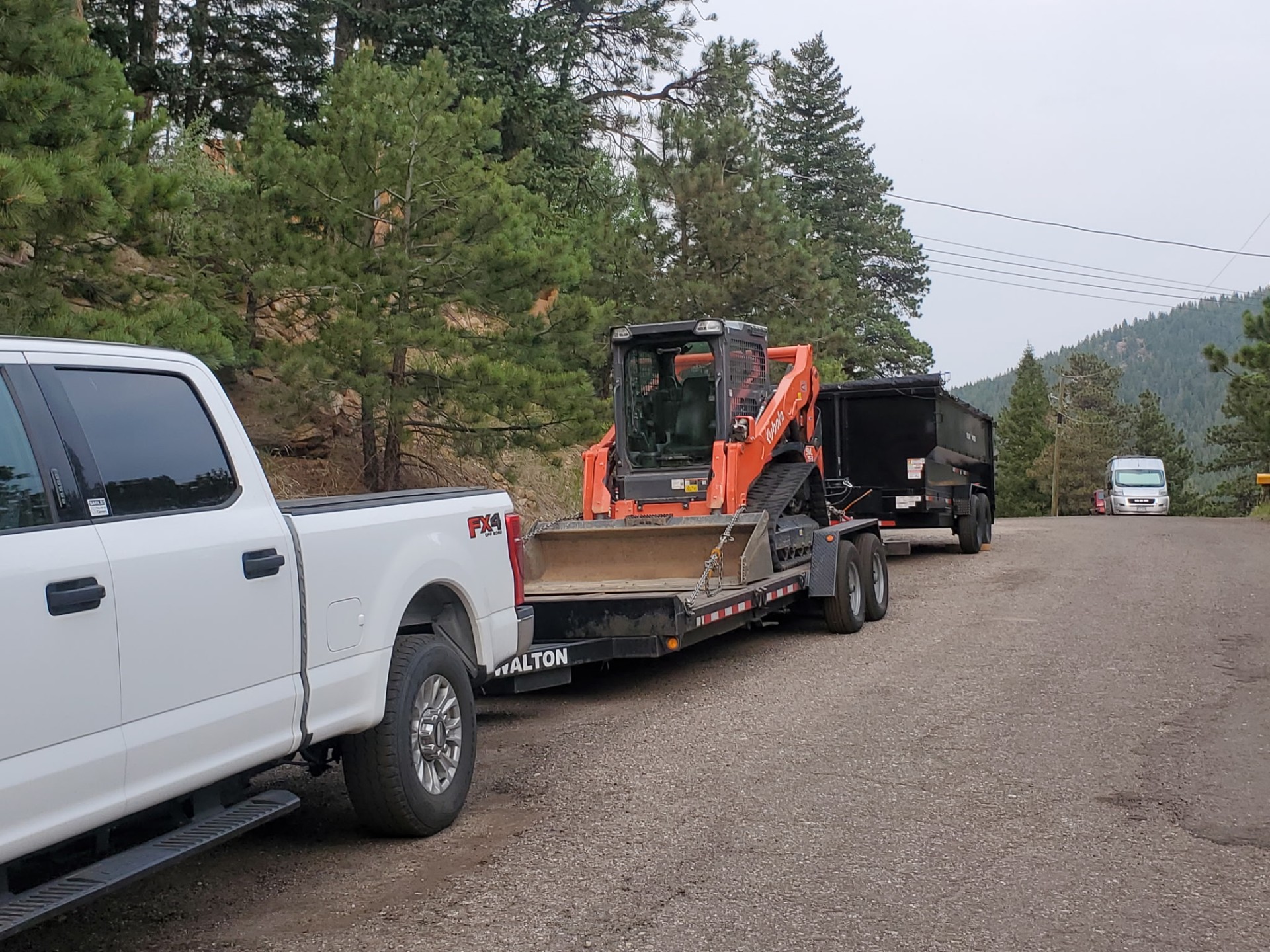 Orange skid steer loading gravel into a black dump trailer with dust flying.