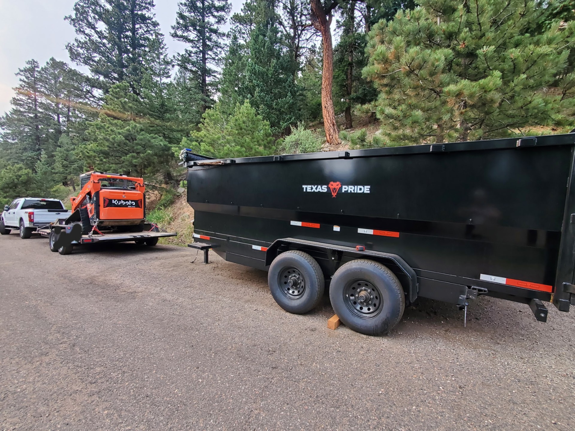 Orange Kubota skid steer with a grapple clearing brush along a wooded tree line.
